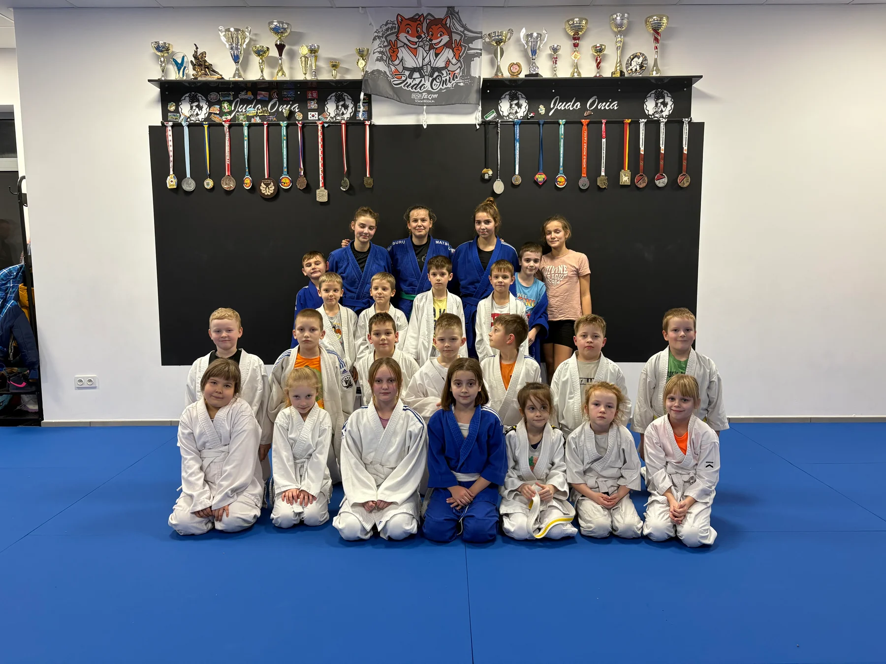 Grupa dzieci i młodzieży Judo Onia na macie pod klubowymi pucharami i medalami